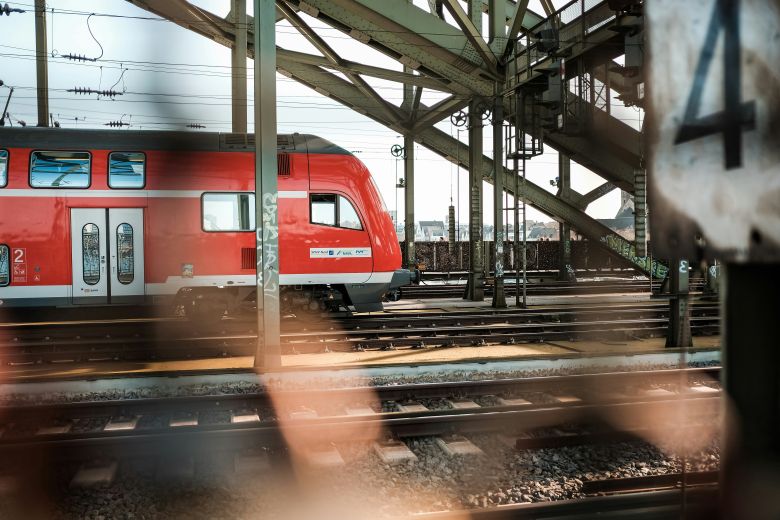 Roter Regionalzug auf einem Gleis unter einer Stahlbrücke, teilweise verdeckt durch unscharfe Reflexionen im Vordergrund.